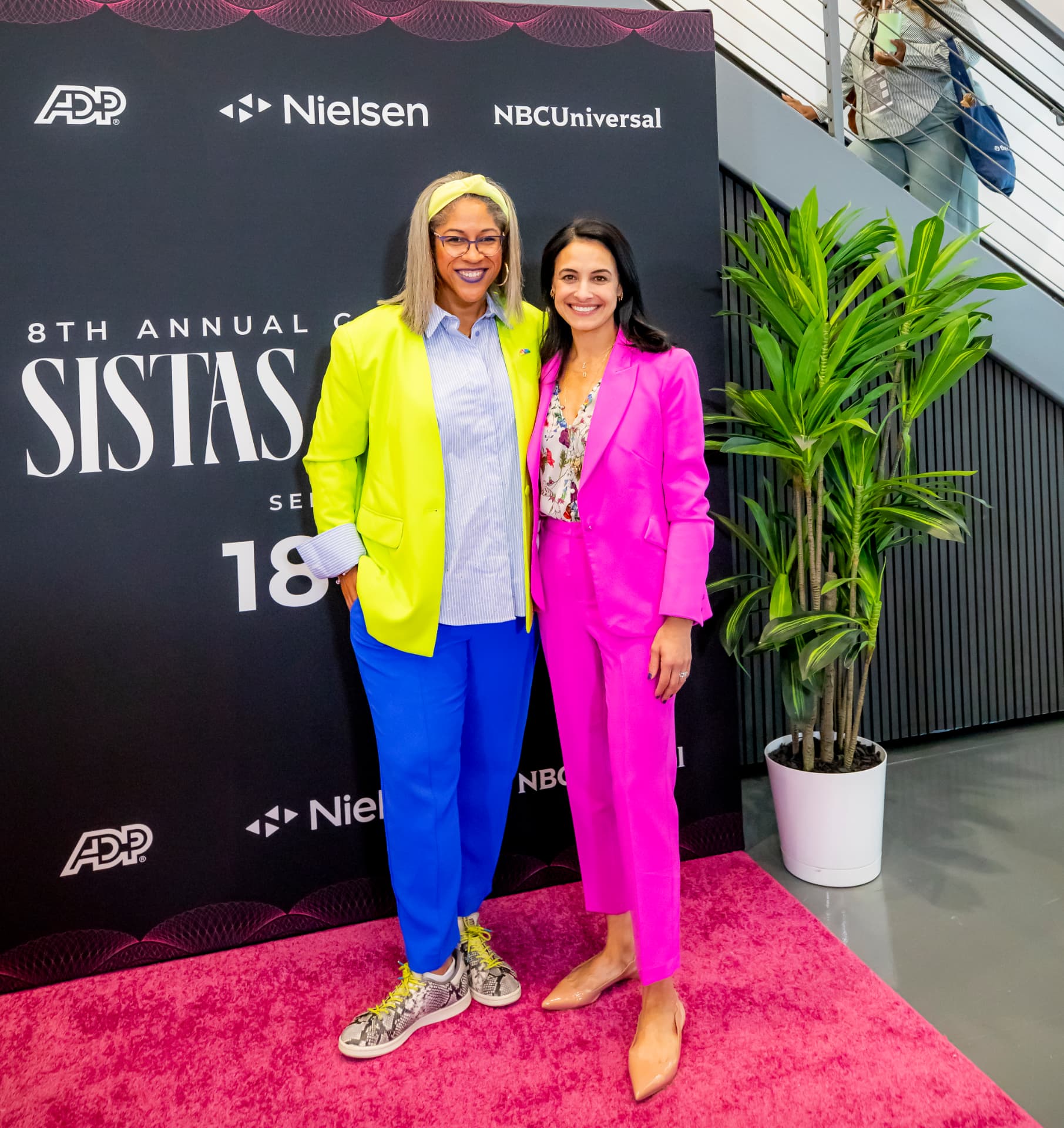 Two attendees on the pink carpet in bold business-chic suits — neon yellow blazer with blue trousers and a hot pink tailored suit