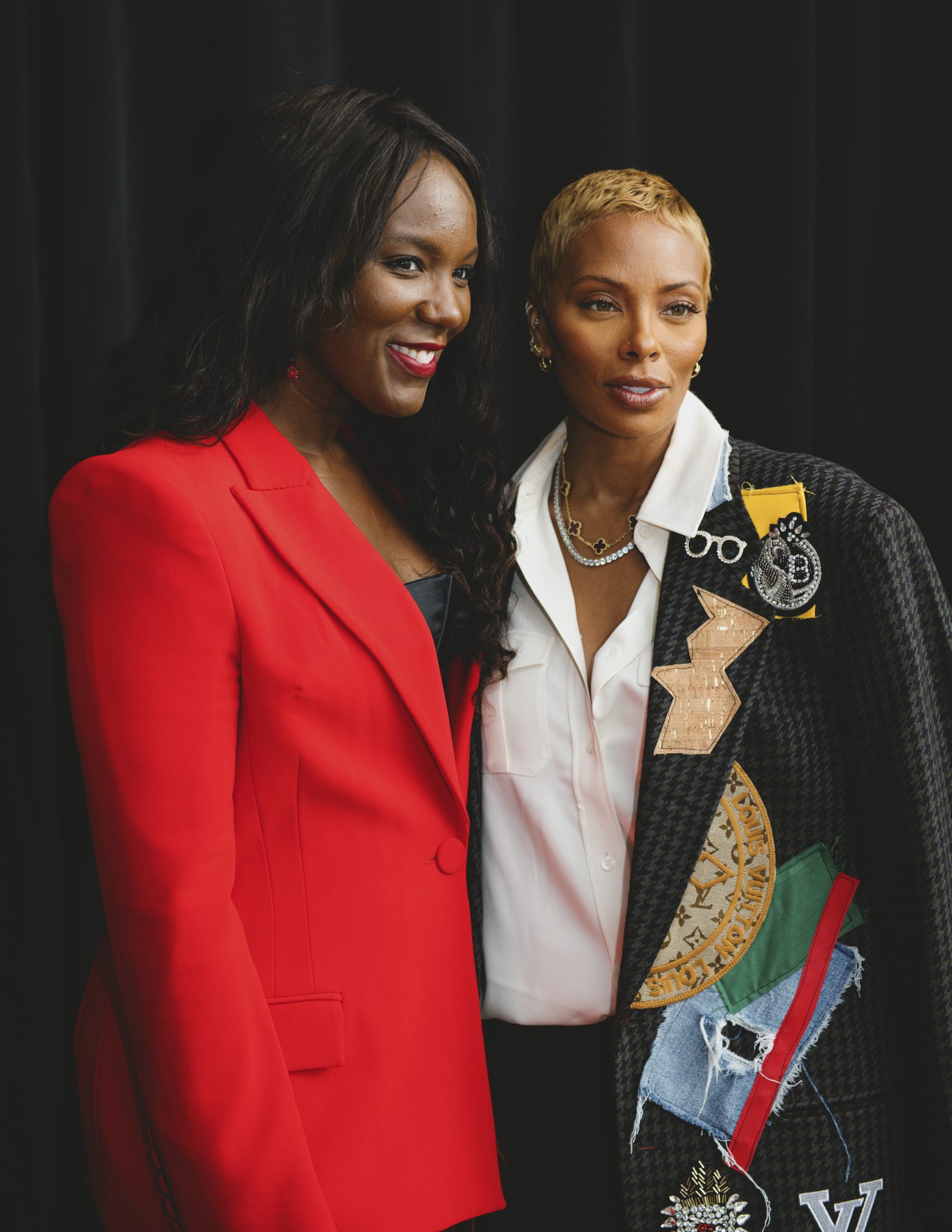 Two women posing together backstage, one in a red suit