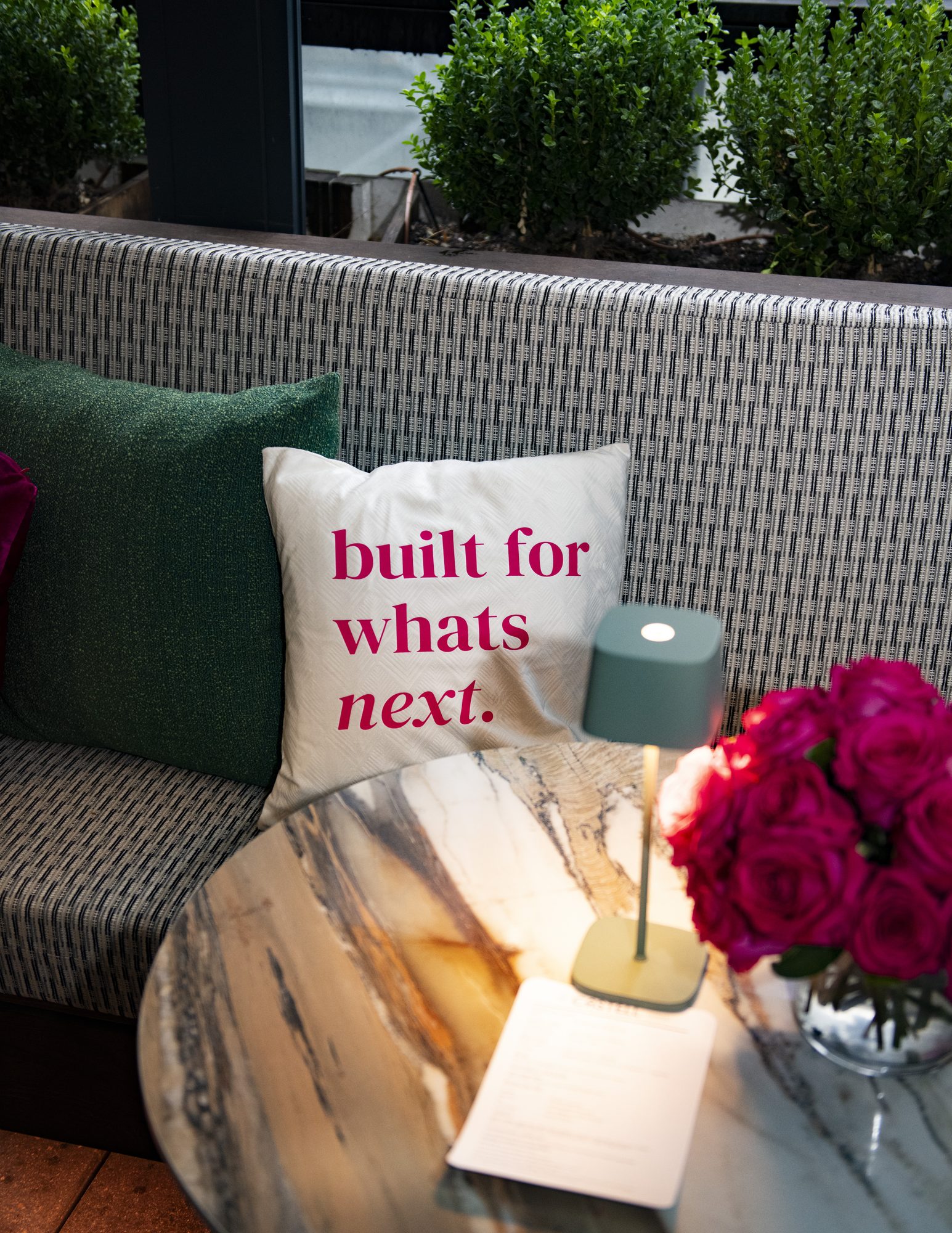 Lounge pillow reading “built for whats next.” beside a table lamp and pink roses